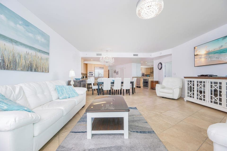 Bright living area featuring a white leather sofas with two queen sleepers, elegant chandelier, and a view into the kitchen