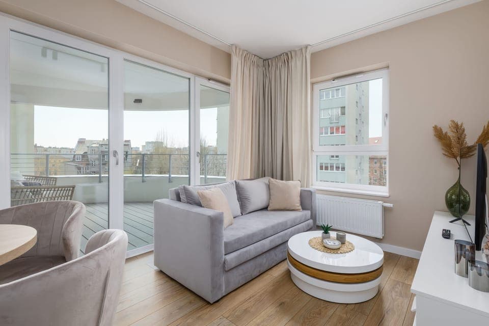 A bright and modern living room with a large glass sliding door leading to a balcony. The room features a round coffee table, a cozy armchair, and neutral-colored decor.