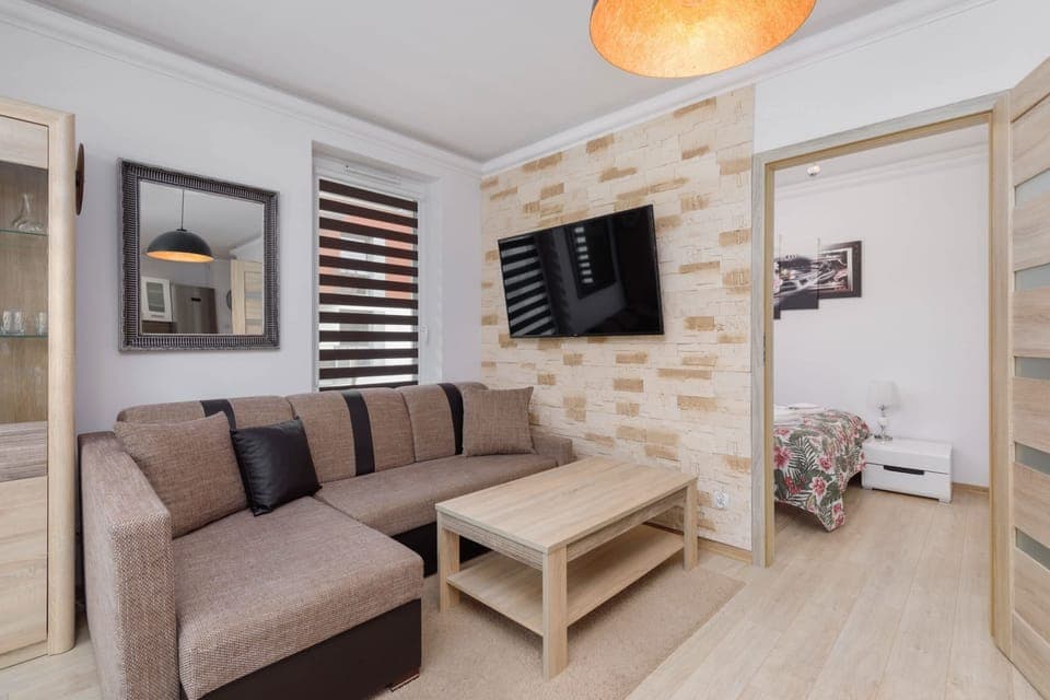 A cozy living room featuring a grey sofa with a wooden coffee table, a flat-screen TV mounted on the wall, and decorative wall panels with a stone-like design.