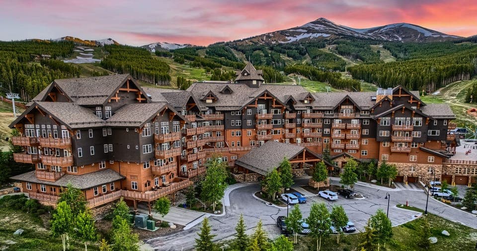 A large rustic mountain lodge with multiple gables and balconies, surrounded by lush greenery and set against a backdrop of forested hills and a pink-hued sunset sky.
