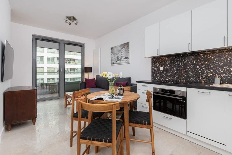 An open-plan kitchen and dining area with white cabinetry, black backsplash tiles, and a wooden dining table for four.

