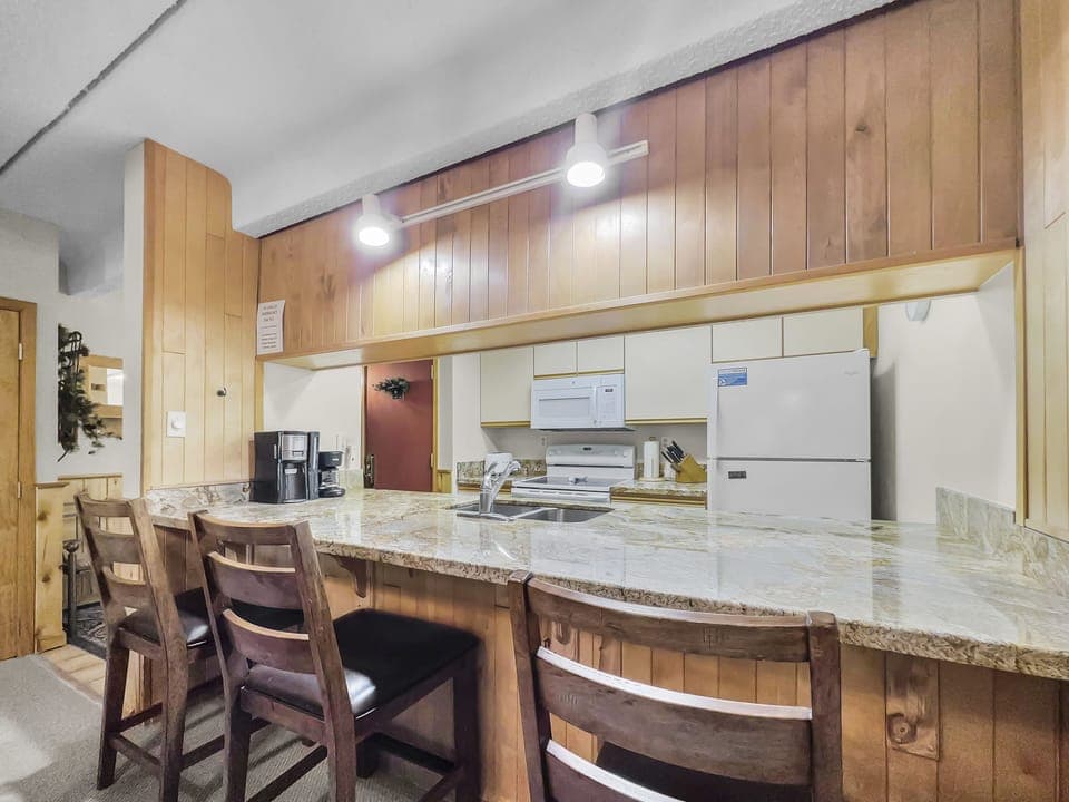 A kitchen with wooden cabinets and a marble countertop, containing three barstools, a coffee maker, a microwave, a stove, and a refrigerator.