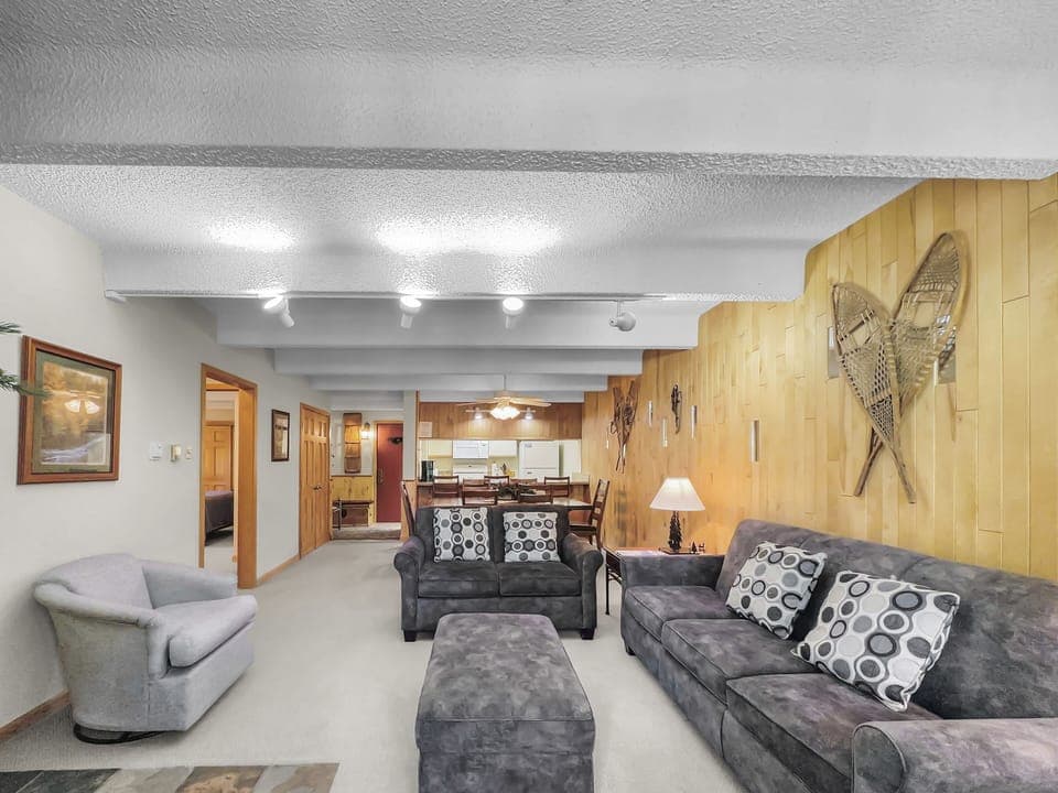 A living room with two couches, an armchair, a coffee table, and framed pictures. The room has wooden paneling, snowshoes as décor, and an open view to the adjacent kitchen and dining area.
