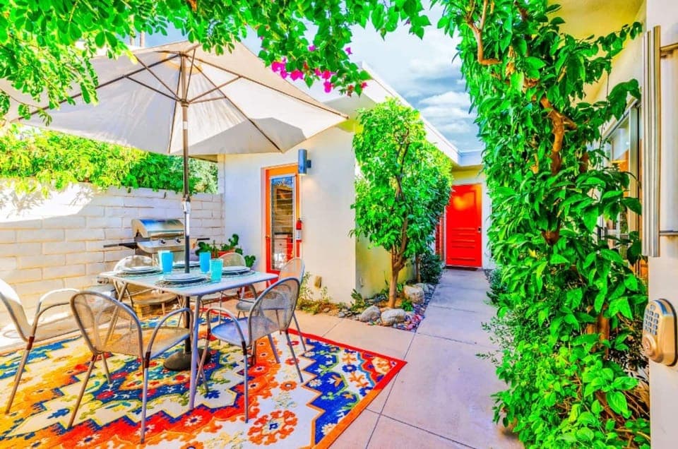 Our patio features a dining set under an umbrella, surrounded by lush greenery, vibrant decor, and bright red doors.