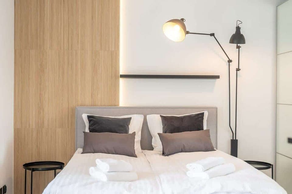 A cozy bedroom with a wooden headboard, white bedding, and black bedside lamps. The room is bright and minimalistic.
