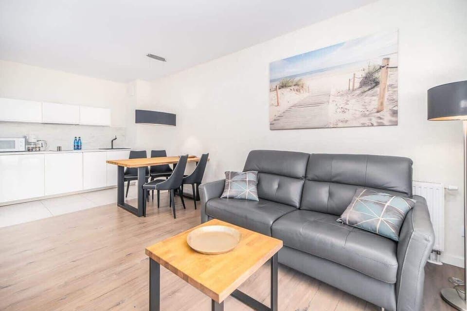 A modern open-plan living area with a gray sofa, wooden coffee table, and a seaside painting, set on light wood flooring.

