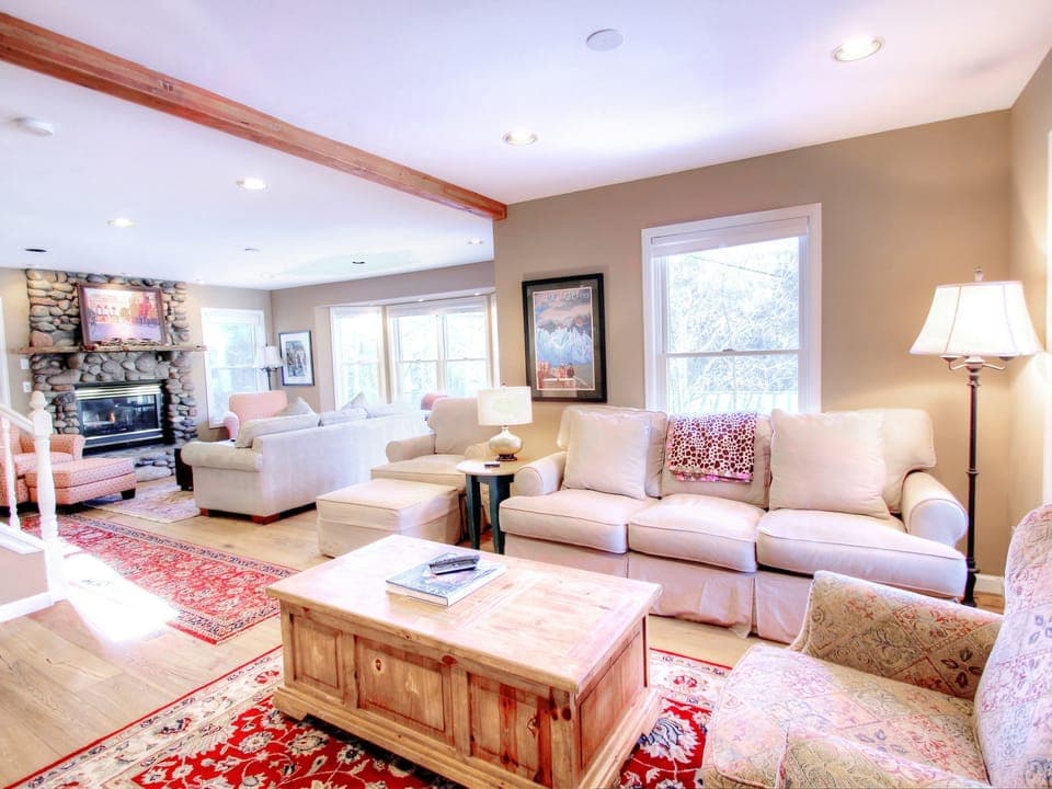 A brightly lit living room with beige sofas, a wooden coffee table, and a stone fireplace. A red rug adds color, and lamps provide additional light. Multiple windows allow natural light to flood the room.