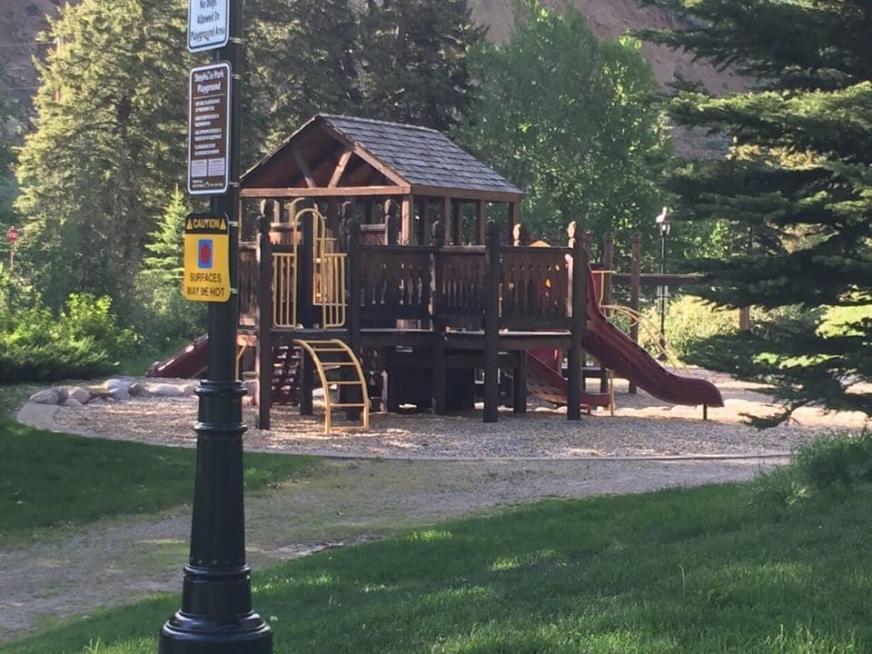 A wooden play structure with slides and climbing features is set in a forested park area. A sign near the structure warns that surfaces may be hot.