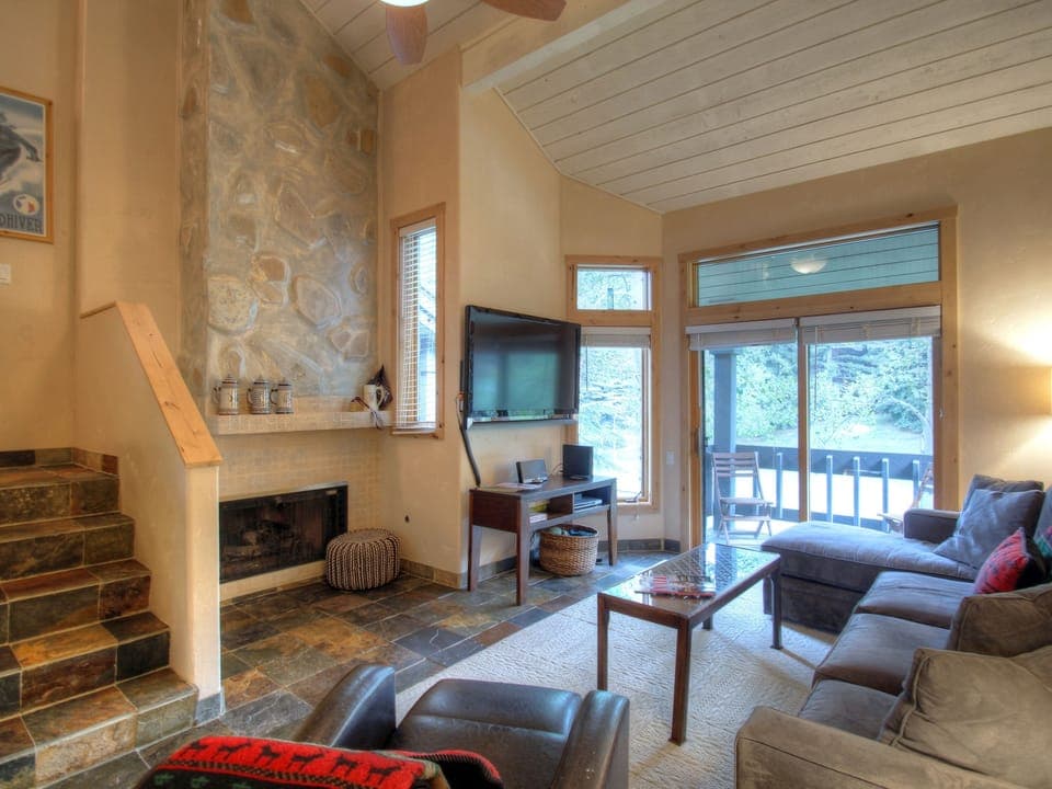 A cozy living room with a stone fireplace, L-shaped sofa, wall-mounted TV, and large windows leading to a balcony. Stairs are on the left, and a lamp illuminates the corner of the room.