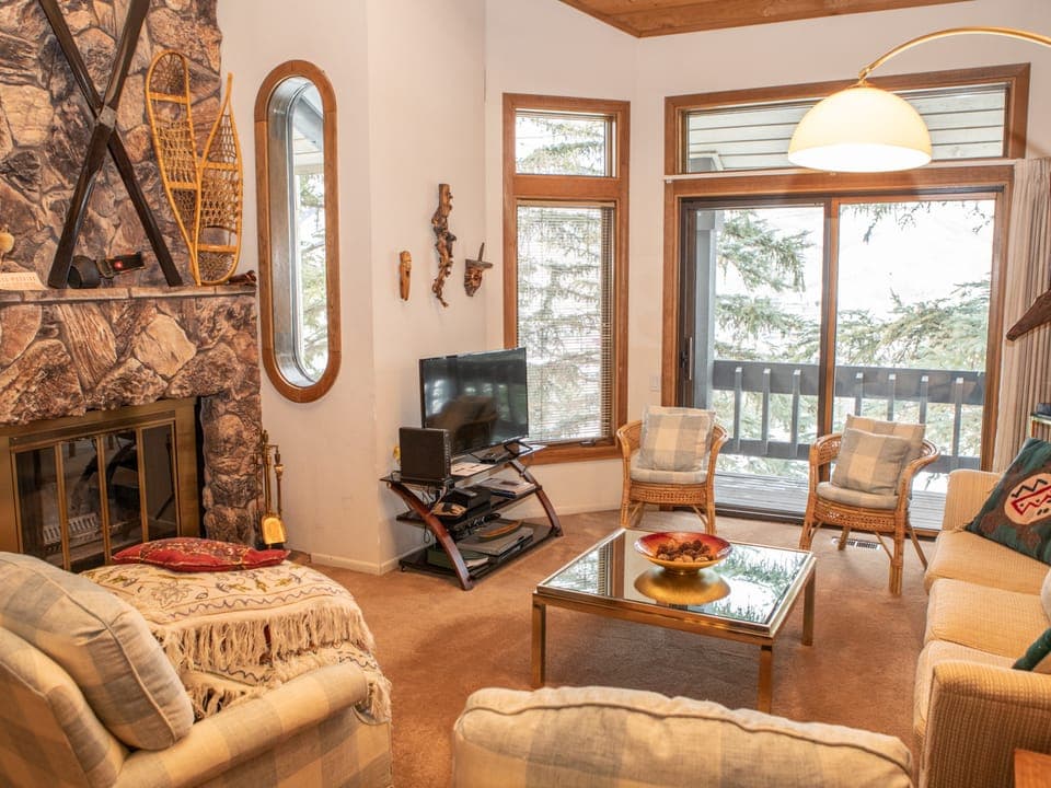 A cozy living room with a stone fireplace, TV, and glass coffee table. Multiple chairs and a sofa provide seating. Large windows and a sliding glass door offer views of a wooded area outside.