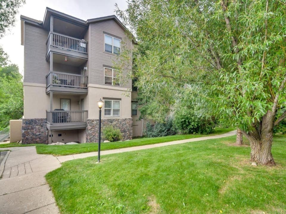 Three-story apartment building with balconies, surrounded by green grass, trees, and a sidewalk. The exterior features beige paint and stone accents. A streetlamp is along the pathway.