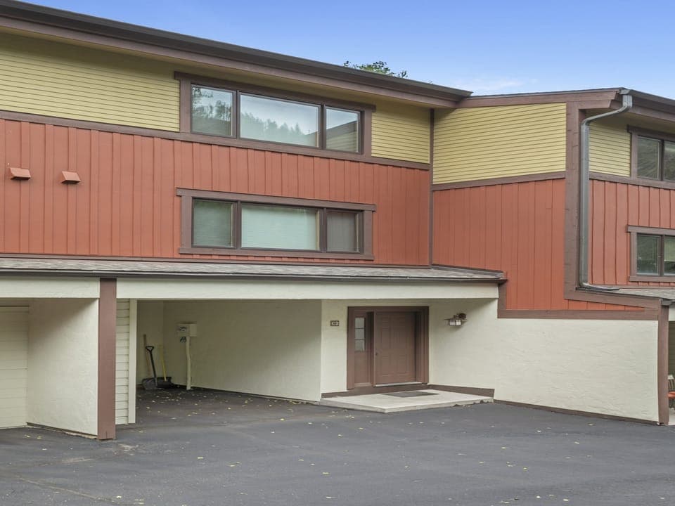 A townhouse with a yellow and red exterior, a brown door, and a covered parking space on a paved driveway.