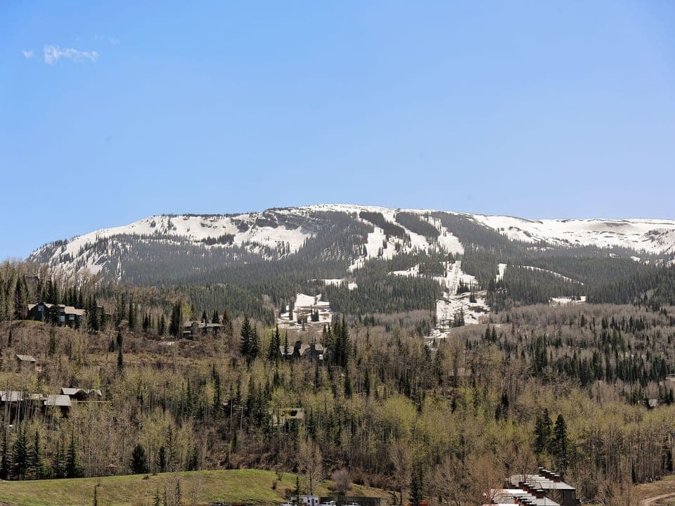 A mountain landscape showing a snow-covered ridge, partially forested slopes, and scattered buildings nestled among trees under a clear blue sky.