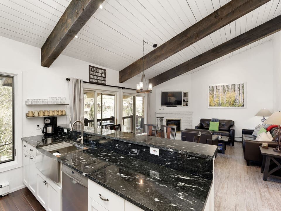 A modern kitchen with a black granite countertop, stainless steel appliances, and an open view into a living area with wooden beams on the ceiling and a fireplace.