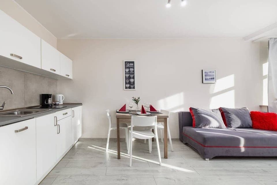 A bright open-plan kitchen and dining area with a white table and a red-accented sofa.

