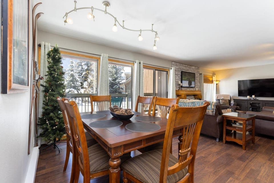 A dining and living area with a wooden table and chairs, a modern light fixture, a TV, a sofa set, large windows, and a holiday tree. The room has hardwood floors and a stone fireplace.