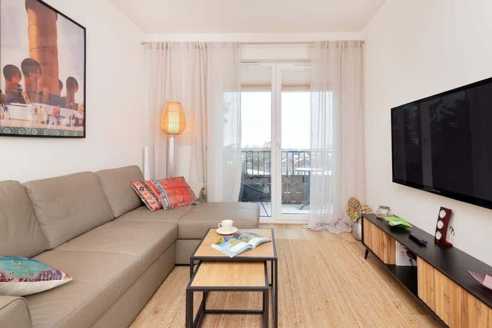 A bright living room with a beige sofa, a wooden coffee table, a TV, and a large window with white curtains.