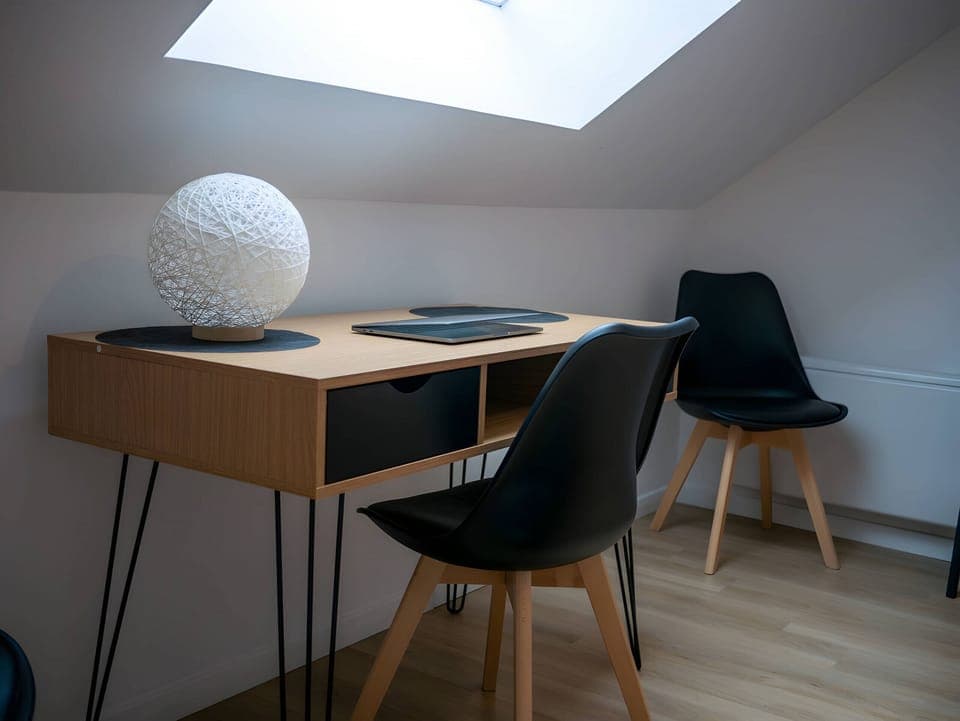 "The wooden desk is placed under a velux, enjoying a pleasant natural light. Its wooden design adds a warm and natural touch to the space. Black chairs are arranged around the desk, offering a comfortable and aesthetic working space.Thanks to the brightness of the velux, this workspace is ideal for stimulating creativity and productivity. The wooden desk and black chairs blend harmoniously to create a welcoming and functional corner to work, study or concentrate. It is an inspiring place to perform your tasks effectively."