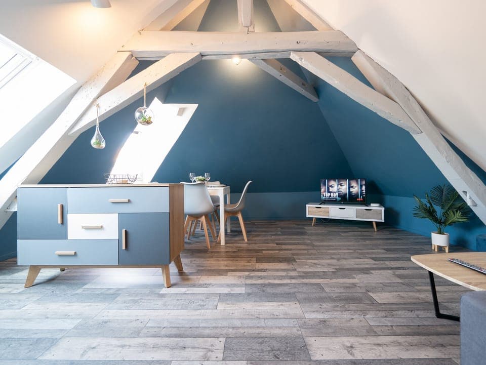 "A space under large attic with white beams, with parquet floor. There is a blue chest of drawers, dining table and well appointed TV area."