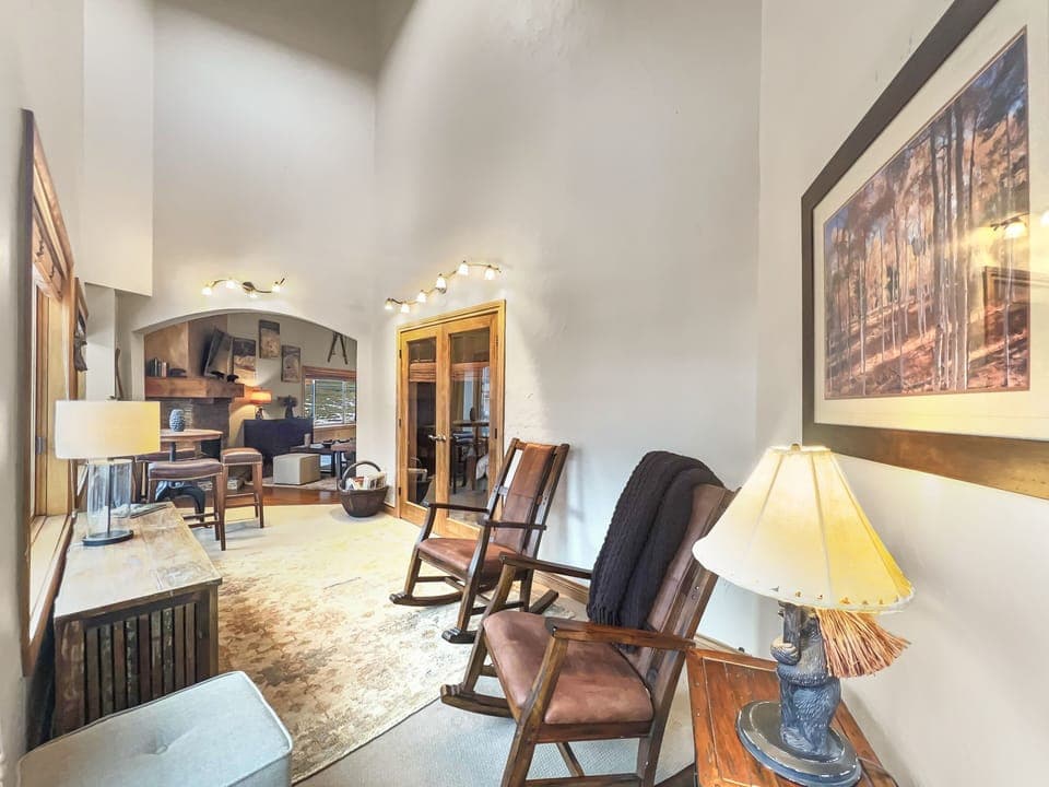 A cozy room features two rocking chairs, a wooden bench, a table lamp, and a framed painting. The room extends to a brighter area with a desk and various furnishings.