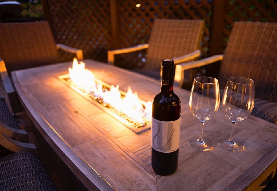 This outdoor space is a perfect evening setting, featuring a rustic wooden table with an integrated fire pit, accompanied by a bottle of wine and glasses.