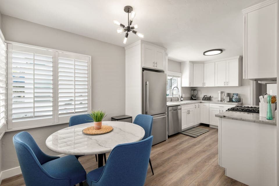 Bright kitchen and dining area with white shaker cabinets, stainless steel appliances, and marble countertops. Modern chandelier hangs over a round marble table with blue chairs. Large windows with shutters fill the space with natural light.