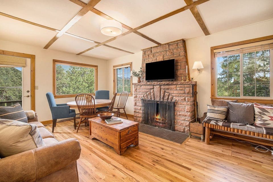 Kittery West Estes Park, Living room with sofa that seats 3 bench coffee table, bench seat with seating for 2, flat screen tv, wood burning fireplace, round dining table with seating for 5, window with view of mountains, and door leading to deck.