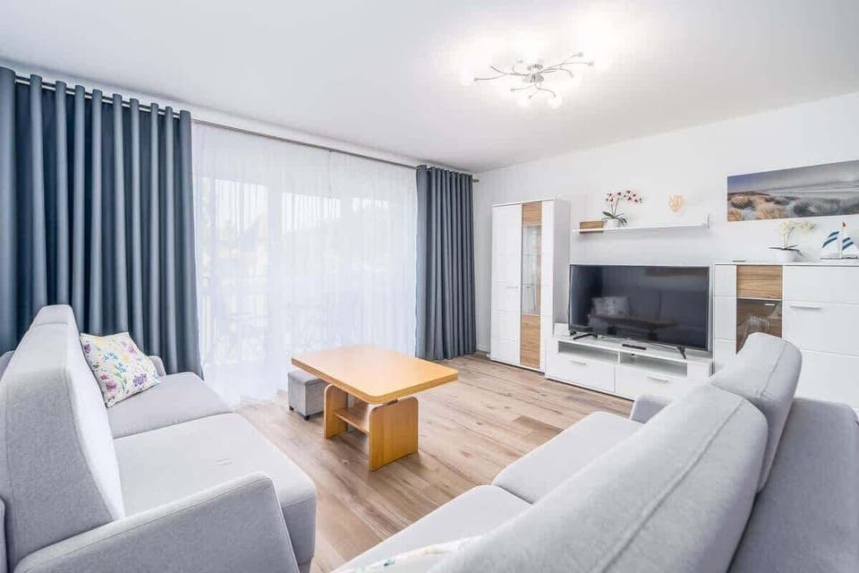 A bright living room with a gray sofa set, a wooden coffee table, and large windows allowing natural light to fill the space.

