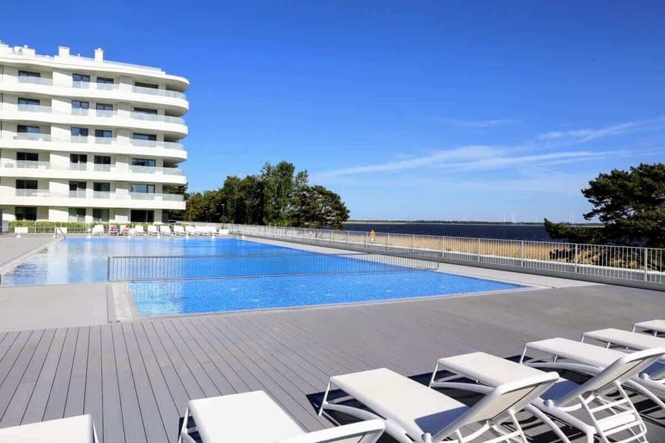 A luxurious outdoor infinity pool with sun loungers, overlooking a modern apartment complex with curved architecture.

