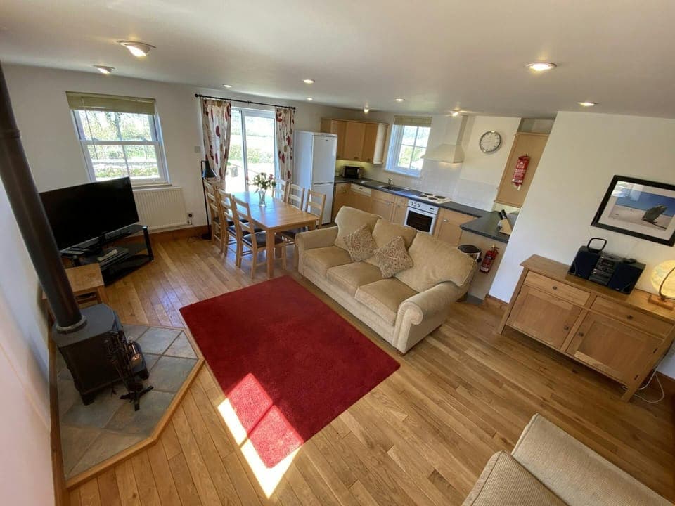 Open plan living/dining/kitchen area with patio doors: sofa, large rug on wooden floor, TV, cabinet with HIFI system and lamp, log burner, dining table with six chairs & well equipped kitchen area