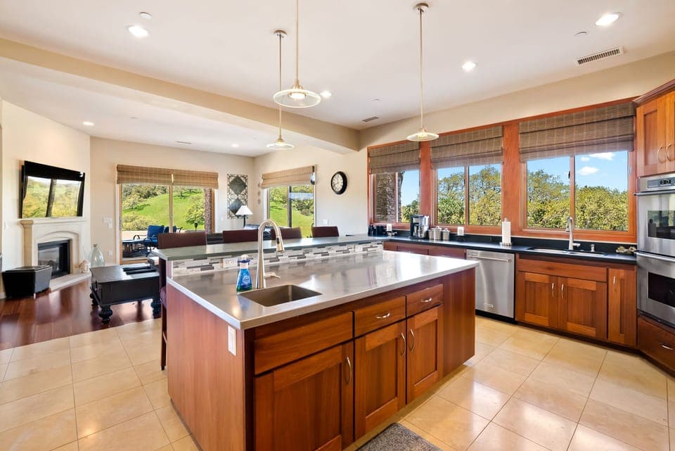 This expansive gourmet kitchen features a massive stainless steel island with a prep sink and custom wood cabinetry. Large windows frame stunning views of the rolling green hills, seamlessly connecting the interior to the breathtaking landscape.