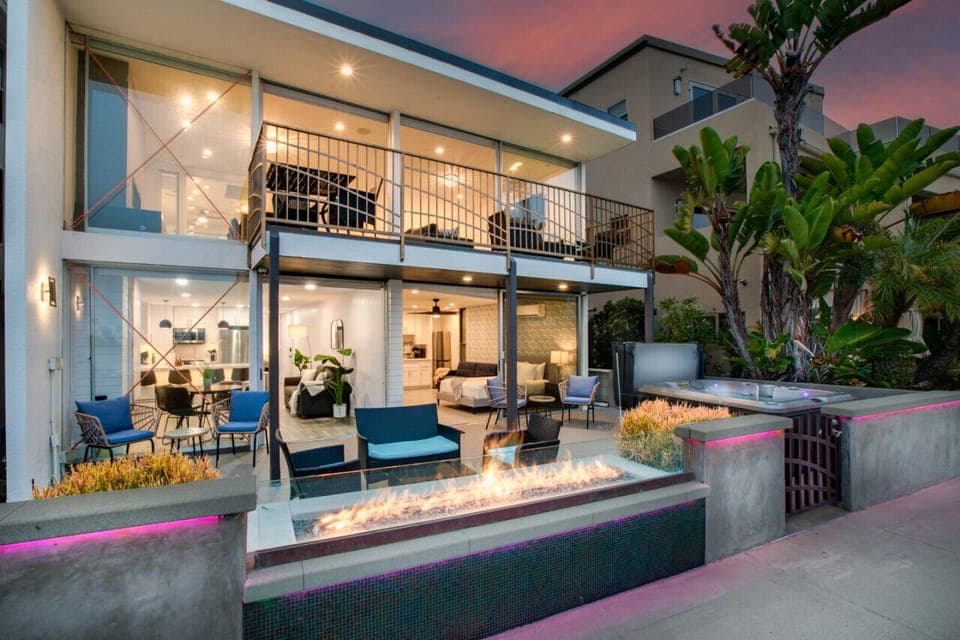 A luxurious beachfront patio at sunset, featuring a cozy outdoor seating area, a modern fire pit, and a shared hot tub. The beautifully lit space seamlessly extends from the interior living area, offering an idyllic setting for relaxation and entertaining