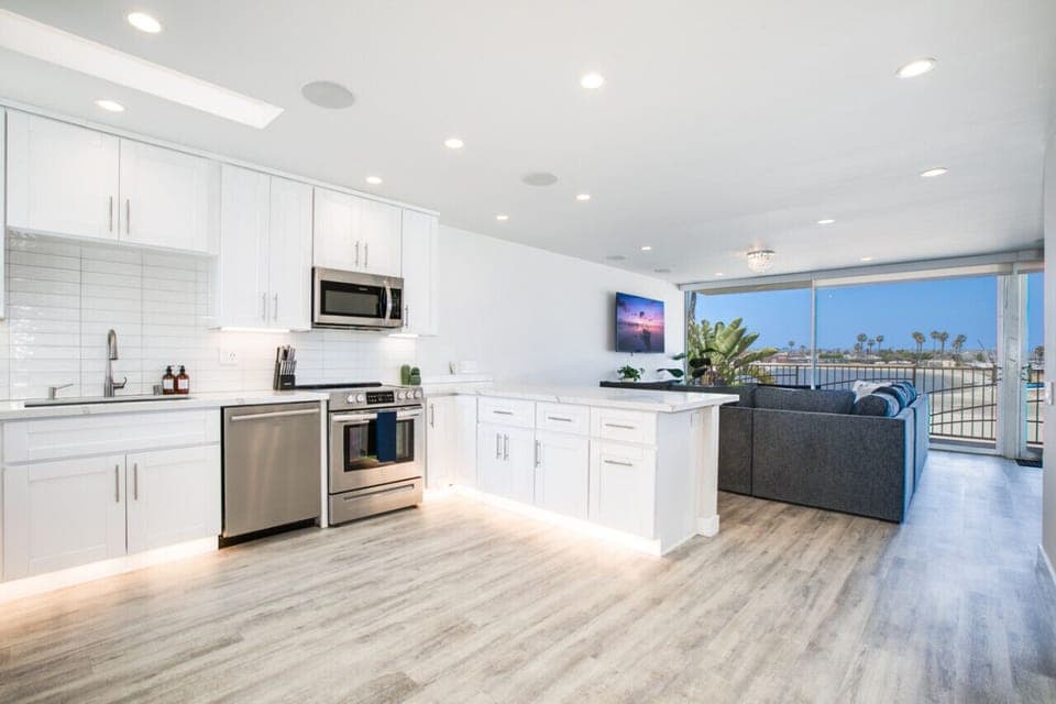 A bright and spacious open-plan living area featuring a modern kitchen with sleek white cabinetry, stainless steel appliances, and under-cabinet lighting. The room seamlessly flows into a comfortable seating area with large windows offering breathtaking v