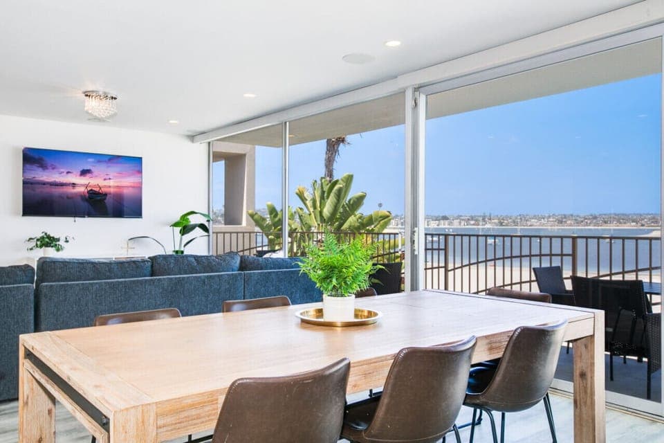 A bright and spacious dining area featuring a large wooden table with modern chairs, illuminated by elegant chandeliers. Floor-to-ceiling windows provide stunning views of the bay and beach, seamlessly connecting the indoor space with the scenic outdoor s