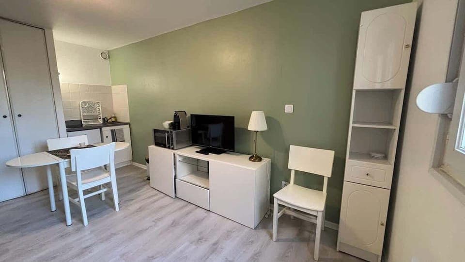 Salon avec canapé convertible et télévision, ouvert sur l'espace de cuisine et séparé de celui-ci par un coin repas pour deux personnes.