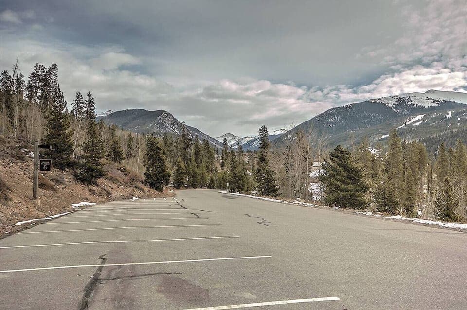 Empty parking lot alongside a forested area with mountain peaks visible in the background under a cloudy sky.