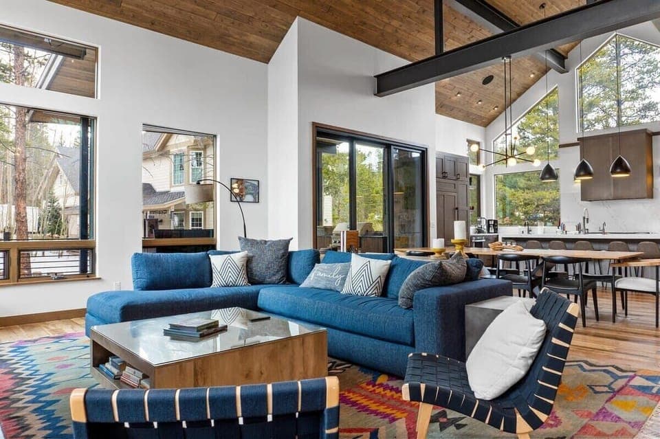 The living room/kitchen/dining room boasts an open and social layout with high-vaulted ceilings and exposed wood beams, soaring floor-to-ceiling windows, and stylish decor.