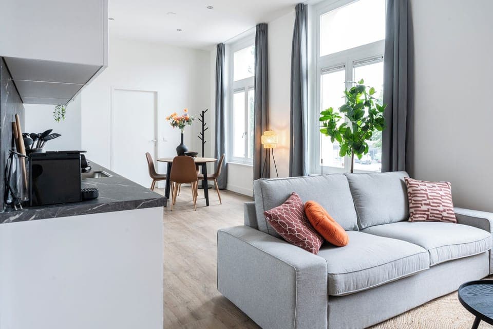 Modern lounge area featuring a cozy gray couch, vibrant orange accents, black round tables, a woven rug, greenery, and tall windows with gray curtains.