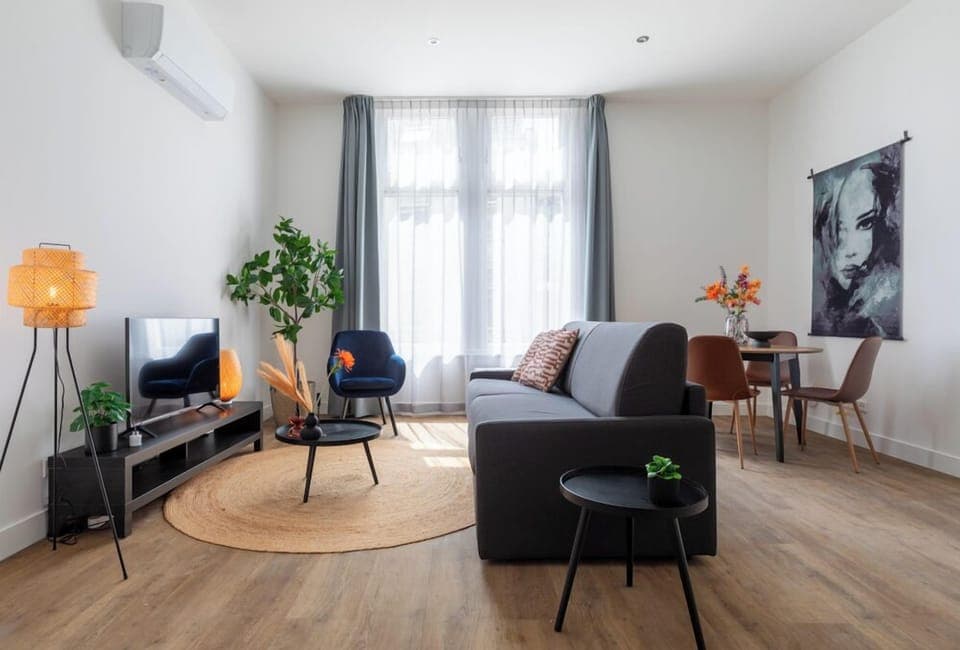 A contemporary living area with a plush sofa, chic chairs, sleek round tables, a TV unit, and natural accents, all enhanced by warm hues and lush greenery, seamlessly linking to the dining space.

Apart-Hotel – close to the university, city center, Efteling & Safaripark Beekse Bergen. Equipped with a new kitchen and a luxury bathroom.