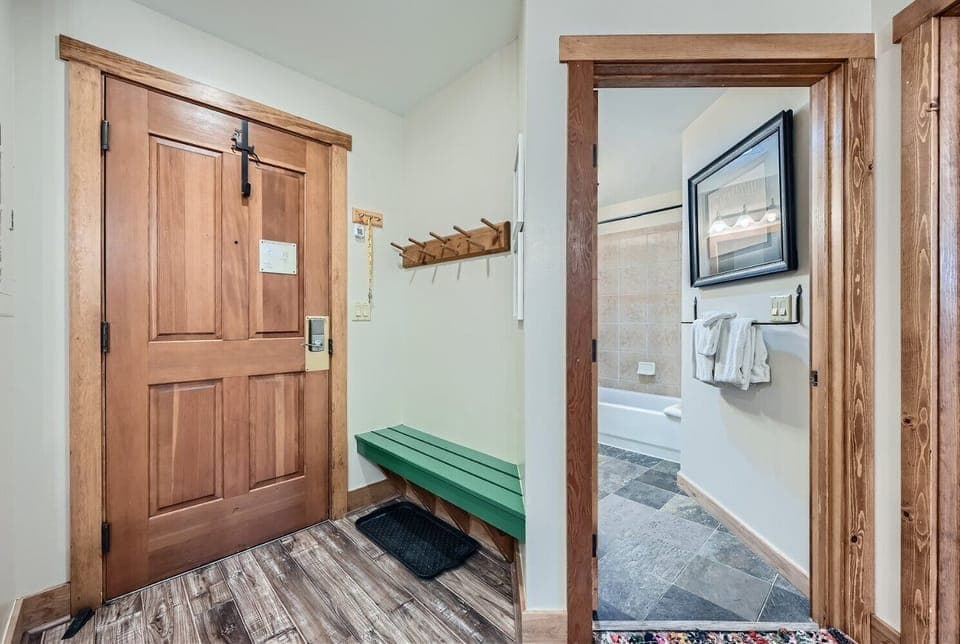 A small entryway with a wooden door, green bench, and coat hooks. Adjacent is a bathroom with a bathtub, tiled walls, and towel rack. The floor has wood and tile finishes.