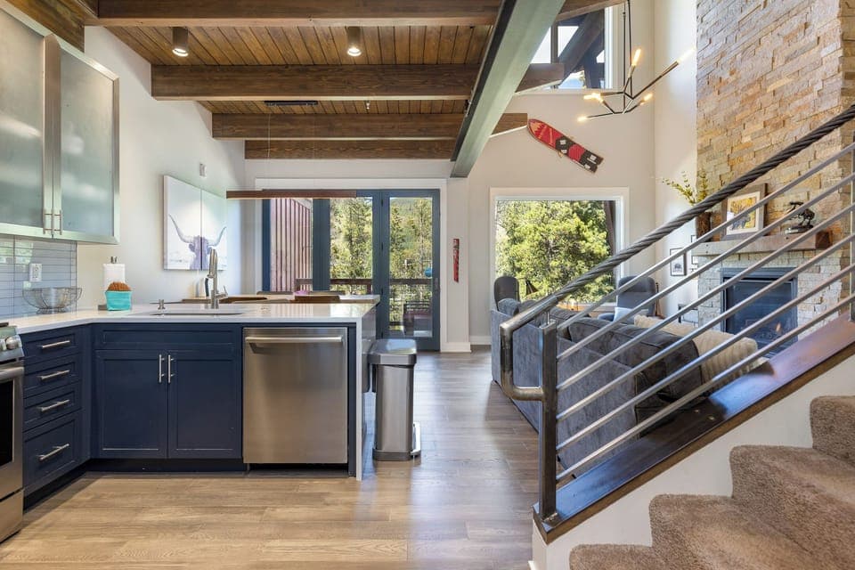 Modern kitchen with dark cabinets, stainless steel appliances, and a wooden beamed ceiling. Large windows and a staircase with a metal railing are visible.