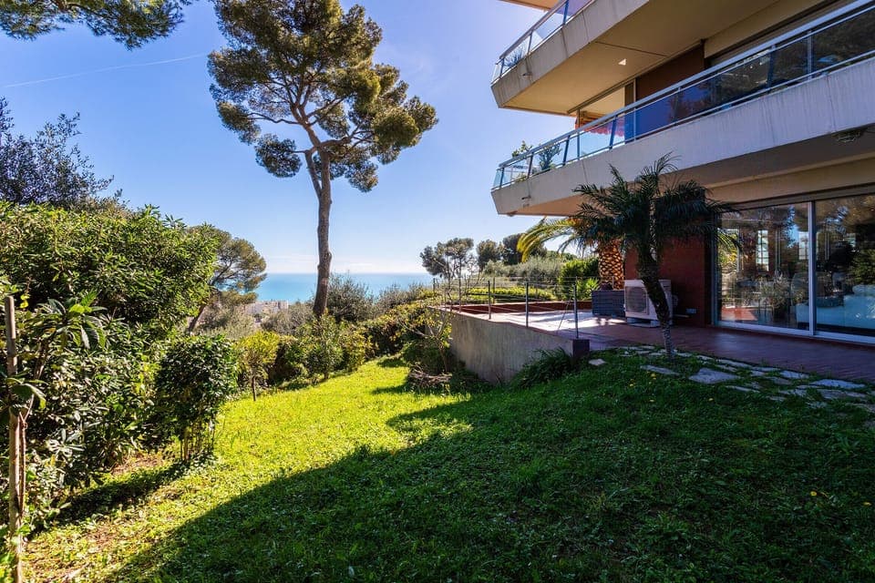 Panoramic view of the sea and city from a lush garden with palm trees and well-maintained hedges.