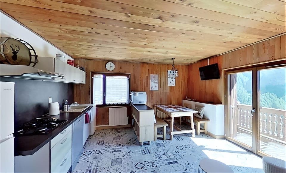 The spacious and well-lit kitchen features modern appliances and ample counter space. The wooden ceiling and walls add a touch of rustic charm. A large window provides natural light, and the dining table with bench seating offers a cozy space for meals. T