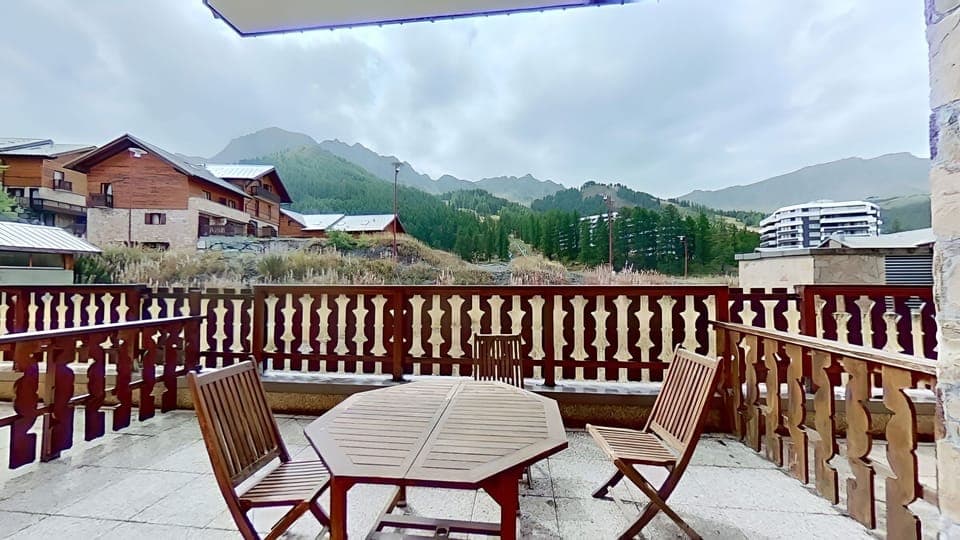 The balcony features a wooden table and chairs, providing a comfortable spot to enjoy the breathtaking mountain views. The surrounding mountains, covered in lush greenery, create a picturesque backdrop. The balcony is enclosed by a wooden railing, ensurin