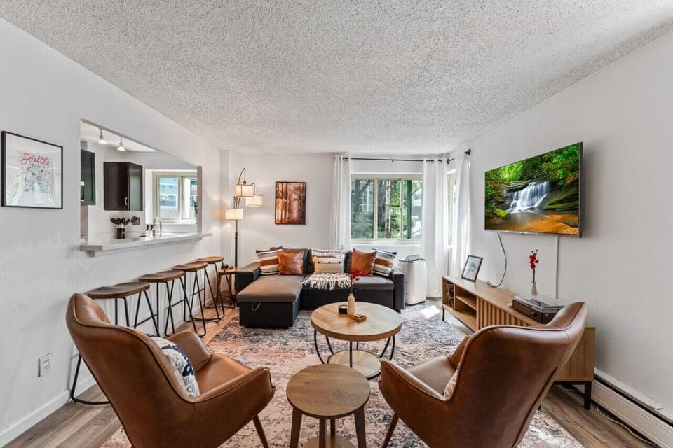 Charming and contemporary living space featuring a sectional sofa, leather chairs, large windows for natural light, open kitchen bar, and a wall-mounted TV with serene views, ideal for a peaceful retreat or social gathering.