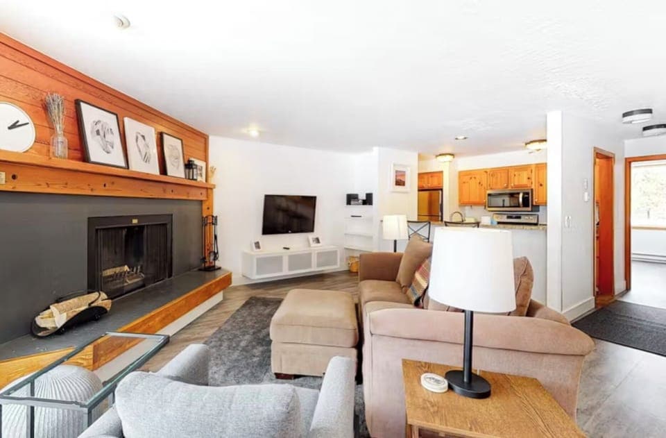 A cozy living room with a fireplace, various artworks above it, a wall-mounted TV, a gray armchair, and a beige sofa. In the background, there's an open kitchen with wooden cabinets and stainless steel appliances.
