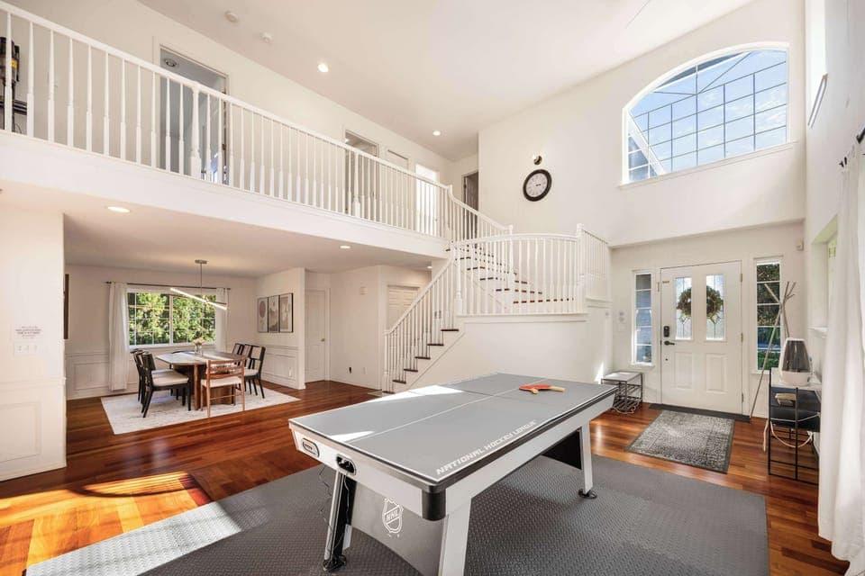 Welcome to our home! Bright, spacious living area with high ceilings, natural light, and a modern air hockey table for entertainment.