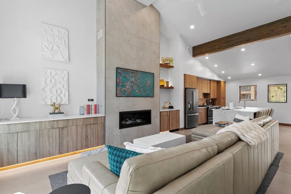 Open-concept living area with vaulted ceilings, mounted TV above a gas fireplace, mini bar with wine fridge, and balcony access.