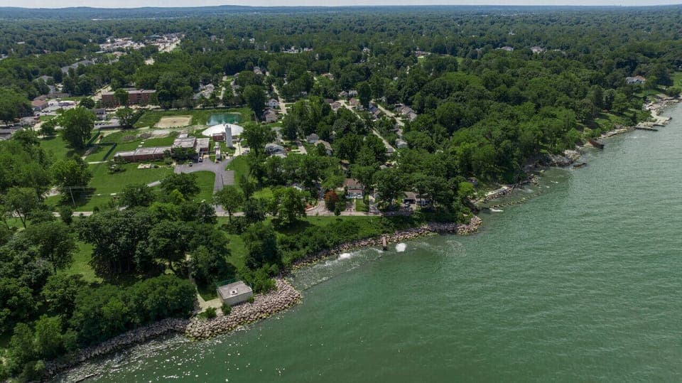 Aerial view showcases the lakefront setting with a lush tree canopy surrounding two homes. The teal house sits beside a landscaped yard, while a neighboring home features a charming red-brick garden. A quiet road curves along the shoreline. 🌳🏡🌊
