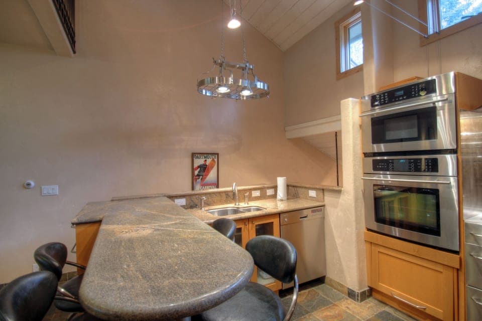 Compact kitchen with a granite countertop bar, stainless steel double oven, sink, and dishwasher. High ceiling with windows, pendant light fixture, stools, and contemporary decor.